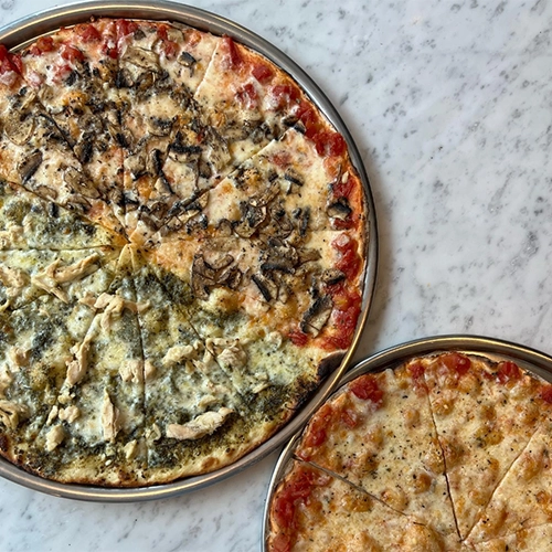 Domatesli, mantarlı ve fesleğen soslu tavuklu olmak üzere üç farklı lezzeti tek bir pizzada buluşturan büyük bir yuvarlak pizza ile klasik domates soslu ve peynirli küçük bir pizzanın beyaz mermer bir yüzeyde, dilimlenmiş ve servis tepsilerinde sunulduğu bu kare, İstanbul'un en iyi pizzacıları listesindeki zengin seçenekleri vurguluyor.