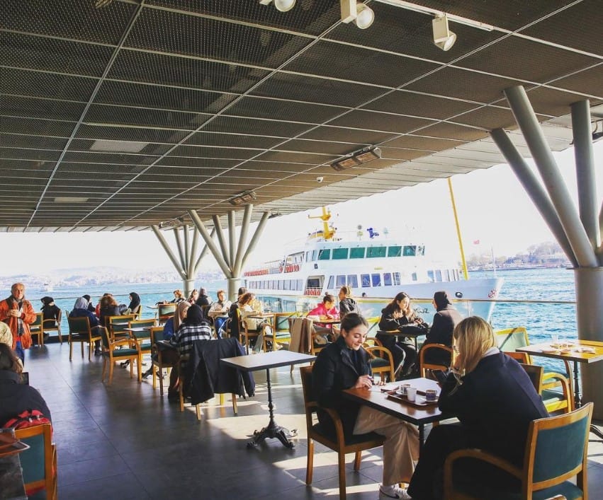 Güneşli bir günde, İstanbul Boğazı'nın eşsiz manzarasına sahip bir teras kafede, insanlar masalarında keyifli vakit geçiriyor; bazıları sohbet ederken, diğerleri sakin bir çalışma ortamı arayanlar için ideal olan bu mekânda işlerine odaklanarak şehrin en iyi çalışma mekânları deneyimini yaşıyor, arka planda bir vapur geçiyor.