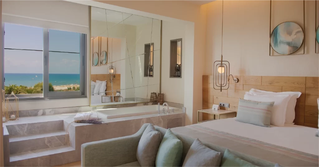 Deniz manzaralı geniş otel odası, büyük yatak ve pencere kenarında modern mermer küvet ile şık bir dekorasyon.