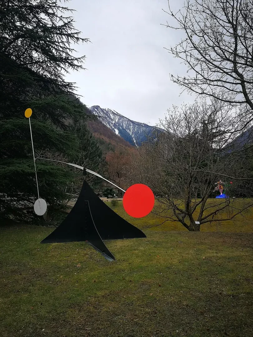 Alexander Calder'ın siyah, üçgenimsi bir stabil form üzerine yerleştirilmiş, uzun çubuklarla dengelenmiş kırmızı, sarı ve gümüş renkli disklerden oluşan mobil parçalara sahip sanat eseri, karlı dağların ve yemyeşil çam ağaçlarının çevrelediği çimenlik bir arazide bulutlu bir havada duruyor.
