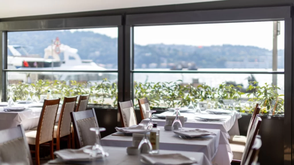 İstanbul Boğazı kıyısında yer alan bir balık restoranının içinden görünen, beyaz masa örtüleri serilmiş masaların özenle kadehler ve tabaklarla hazırlandığı, dışarıda yemyeşil bitkilerin ve uzaklarda geçen gemilerin Boğaz manzarasına eşlik ettiği ferah ve aydınlık bir ambiyans.