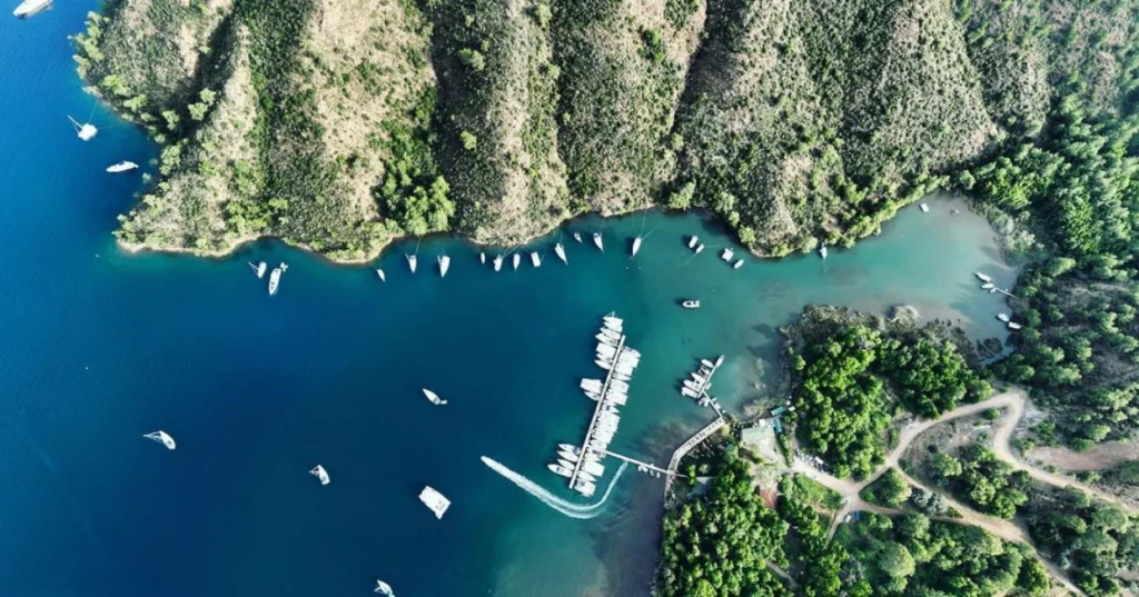 Yeşil tepeler ve yemyeşil ormanlarla çevrili, Göcek'in berrak turkuaz sularında yüzen çok sayıda yelkenli ve teknenin demirlediği Biynuzbükü koyunun huzurlu bir kuşbakışı görüntüsü.