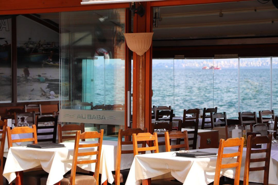 Büyükada'da yer alan Ali Baba restoranının iç mekanından çekilen bu fotoğrafta, deniz manzaralı camların önünde boş bekleyen beyaz örtülü masalar ve ahşap sandalyeler, uzaktaki sakin denizi ve bir teknenin siluetini gözler önüne seriyor.