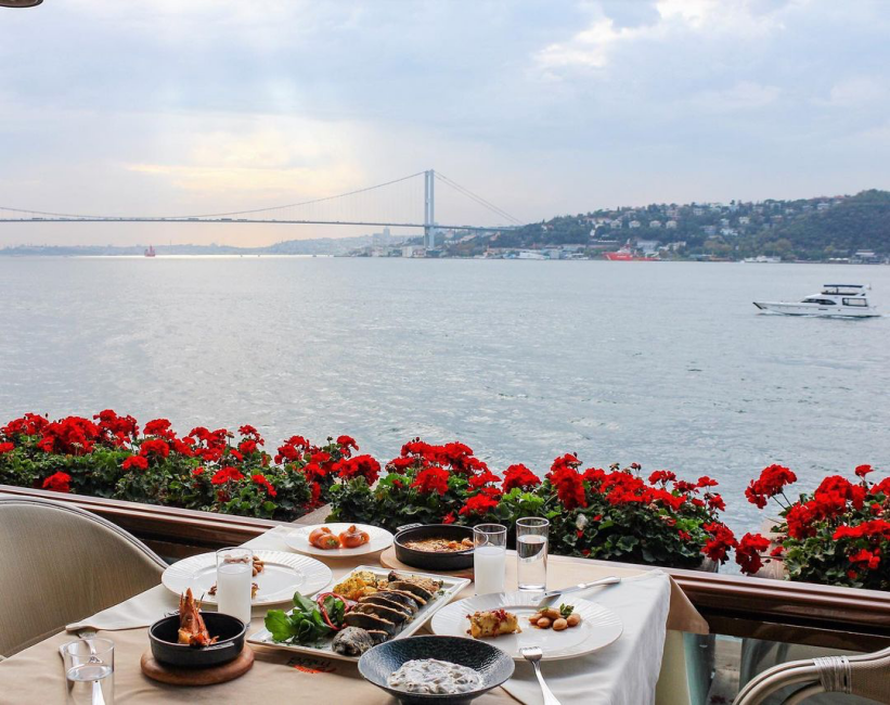 İstanbul Boğazı'nın huzurlu sularına ve uzaklardaki köprüye nazır, kırmızı çiçeklerle süslü bir terasta, taze balık, karides ve geleneksel mezelerle dolu zarif bir yemek masası gökyüzünün gri tonlarıyla bütünleşiyor.