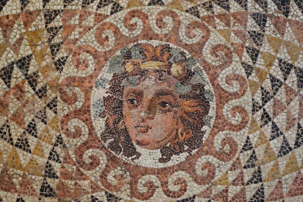 Antik bir mozaiğin merkezinde, yaprak ve sarmaşıklarla süslenmiş bir çelenk taşıyan, kahverengi tonlarında bukleli saçlara ve altın rengi küpeye sahip genç bir yüz, ayrıntılı mozaik taşlarıyla özenle işlenmiş; etrafı kahverengi ve beyaz sarmal desenlerle, dış kısımları ise üçgen geometrik motiflerle çevrilidir.