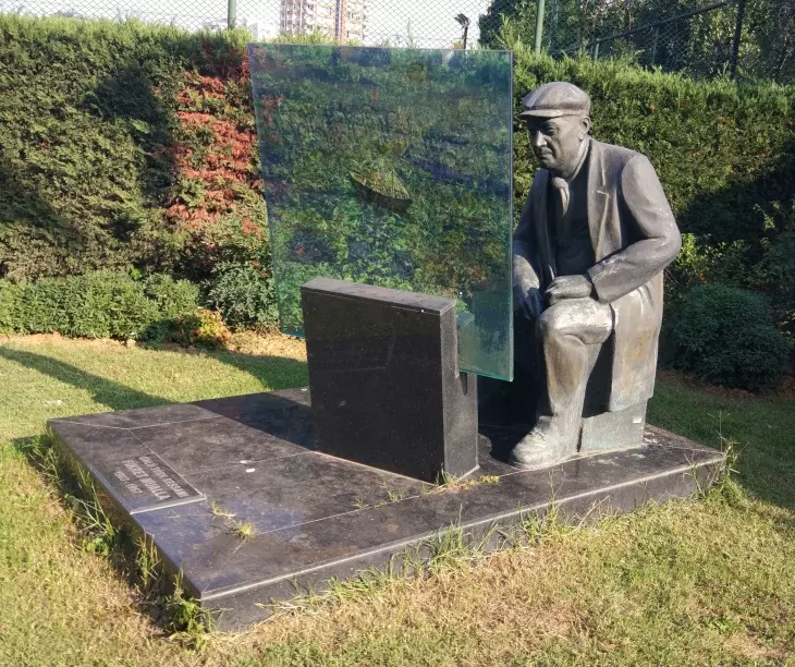 Fikret Mualla'nın bronzdan yapılmış heykeli. Oturan pozisyondaki heykeltıraş, yanında bir cam levha ve bir mezar taşı detayıyla, sanatçının anıt mezarının veya bir anma heykelinin bulunduğu açık hava alanını gösteriyor. Heykelin arkasında yeşil ağaçlar ve bir çit bulunuyor.