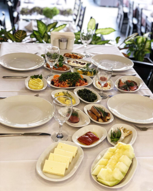 Beyaz masa örtülü bir restoranda, ortasında bol yeşillikli bir salata ile tereyağı, dilimlenmiş kavun ve kırmızı biber gibi çok sayıda farklı soğuk mezenin küçük beyaz tabaklarda özenle dizildiği, boş yemek tabağı ve kadehlerin eşlik ettiği zengin bir sofra gözüküyor.