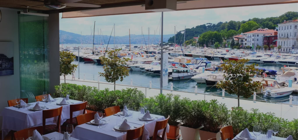 Şık beyaz örtülü masaların ve sandalyelerin sıralandığı, yarı açık bir restoranın ön planında, masmavi sularla dolu bir marinada demirlemiş çok sayıda lüks tekne ve yatın, yemyeşil tepelerin ve kıyıdaki binaların oluşturduğu büyüleyici bir manzara uzanıyor, deniz kenarında keyifli bir yemek deneyimi için davetkar bir atmosfer sunuyor.