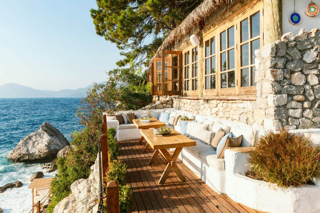 Deniz kenarında kayalıkların üzerine kurulmuş, taş duvarlı ve ahşap pencereli Nautical Hotel restoran terası, ahşap masalar ve deniz manzarasıyla romantik bir atmosfer sunuyor.
