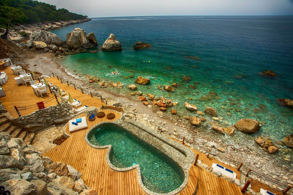 Nautical Hotel’in kayalıklar arasındaki sahilinde turkuaz deniz manzarasına karşı ahşap iskele, oturma alanları ve taş havuz bulunuyor.