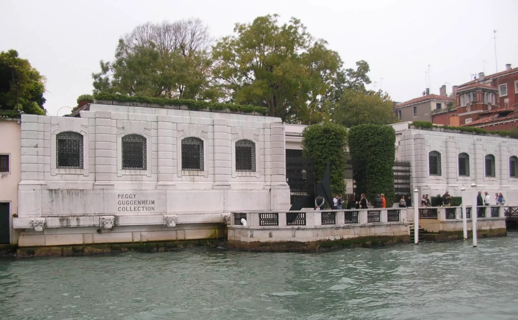 Venedik'teki Peggy Guggenheim Koleksiyonu müzesi, gri bir gökyüzünün altında yeşil bir kanalın kıyısında yer alan, cephesinde PEGGY GUGGENHEIM COLLECTION yazan, demir parmaklıklı pencereleri ve önünde dolaşan ziyaretçileri olan beyaz taş bir yapıdır.
