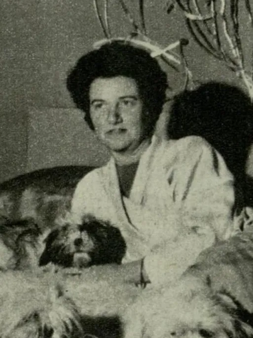 Tanecikli siyah beyaz bir fotoğrafta, koyu kıvırcık saçlı ve açık renk bir giysi giyen Peggy Guggenheim, doğrudan kameraya bakarken, etrafı çeşitli küçük, tüylü köpeklerle çevrili bir şekilde, kişisel alanındaki sakin anlarından birini yansıtıyor.