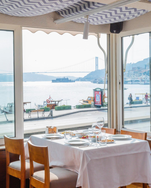 Beyaz örtülü, mezelerle donatılmış bir masanın ve ahşap sandalyelerin bulunduğu ferah bir restorandan Bosphorus Köprüsü'nün ve hareketli sahilin parlak gün ışığıyla aydınlatıldığı manzarayı gösteren, pencereden dışarıya bakan bir görünüm.