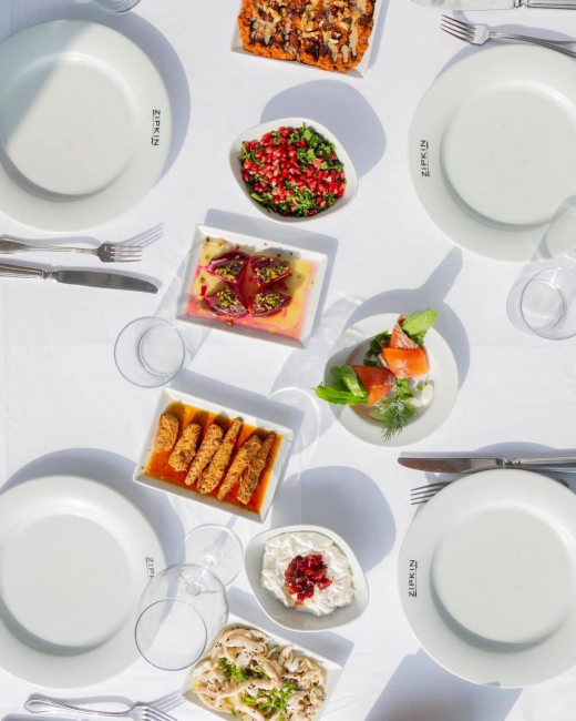 Beyaz örtülü bir masada yukarıdan çekilmiş bu görüntüde, ZİPKİN logolu boş tabaklar, nar taneli yeşil salata, somon füme ruloları, mantar salatası, kırmızı soslu kızarmış deniz mahsulleri ve fıstıklı pembe mezeler dahil zengin çeşitlilikte Ege ve Akdeniz mutfağına özgü başlangıçlar sergileniyor.