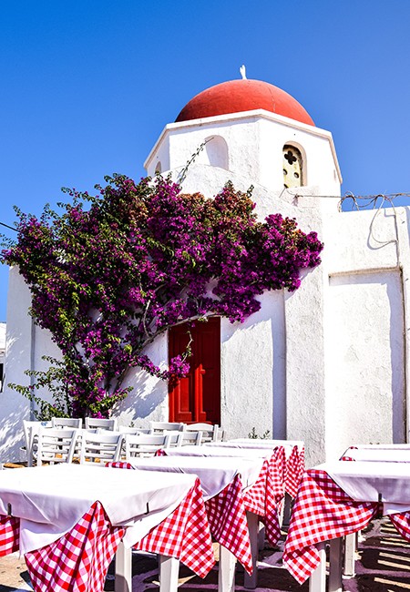 Paros'un karakteristik beyaz badanalı binalarından birinin önünde, kırmızı kubbeli bir yapıyı süsleyen mor begonviller ve kırmızı beyaz kareli masa örtüleriyle dolu, sıcak bir Yunan Adası tavernasından canlı bir an.