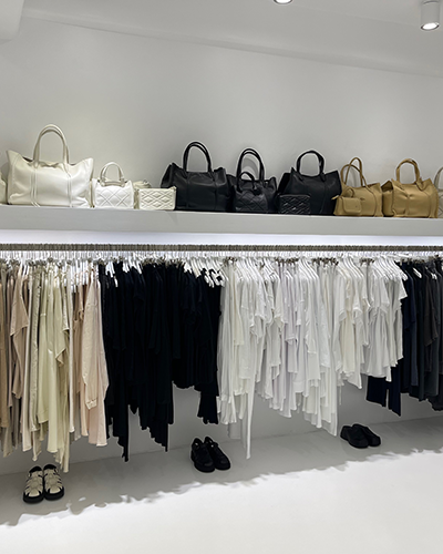Paros seyahat rehberi bağlamında Angel's mağazasından bir görünüm: Beyaz ve minimalist iç mekanda, üst rafta beyaz, siyah ve ten rengi Callista çantaları şıkça dizilmiş, alt kısımdaki askılıklarda ise bej, siyah, beyaz ve gri tonlarında lüks bohem kıyafetler sergileniyor; zeminde ise iki çift ayakkabı görülüyor.
