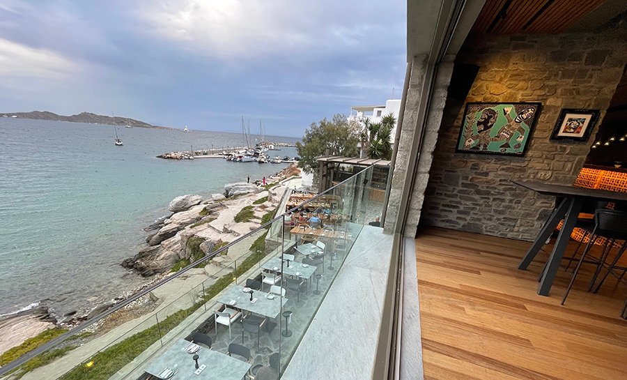 Paros Adası'nda, deniz kenarındaki modern Deck Paros restoranından, berrak turkuaz sulara, kayalık kıyıya ve teknelerin demirlediği Naousa limanına bakan, cam korkuluklu açık hava yemek alanını ve iç mekandaki taş duvar detaylarını gösteren genel bir görünüm, Paros seyahatini keşfetmek için cazip bir atmosfer sunuyor.