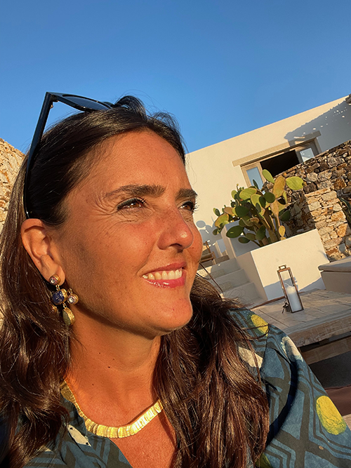 Paros seyahat rehberi için yazan Deniz Yıldız Edin, güneşin aydınlattığı bronz teni ve koyu renk saçlarıyla, başında güneş gözlüğü ve boynunda altın kolyesiyle, arka planda Paros'un tipik beyaz ve taş duvarlı bir yapısının ve bir kaktüsün bulunduğu huzurlu bir Akdeniz ortamında gülümseyerek uzaklara bakıyor.