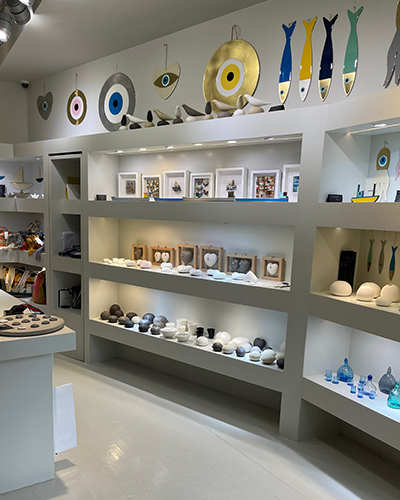 Paros'taki Hera's adlı sanat ve el işi mağazasının içinden bir görünüm; beyaz raflarda seramik balıklar, nazar boncuğu temalı duvar süsleri, mermer kalpler ve çeşitli ev dekorasyon ürünleri Paros seyahat rehberi okuyucularına özgün alışveriş noktalarını tanıtıyor.
