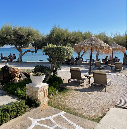 Paros'un sakin atmosferini yansıtan bu sahil manzarası, konforlu şezlonglar ve şemsiyelerin bulunduğu çakıllı bir plajda gölgelik ağaçların altında dinlenen insanlarla dolu olup keyifli bir Paros seyahat deneyimini vurguluyor.