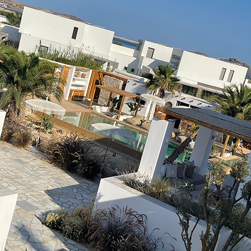 Paros'taki çok yeni ve bohem-şık bir konaklama alternatifi olan Kosmitis Suits'in bembeyaz modern yapıları, palmiyelerle çevrili açık havuzları ve şık dış mekan oturma alanlarını gösteren güneşli bir genel görünüm.