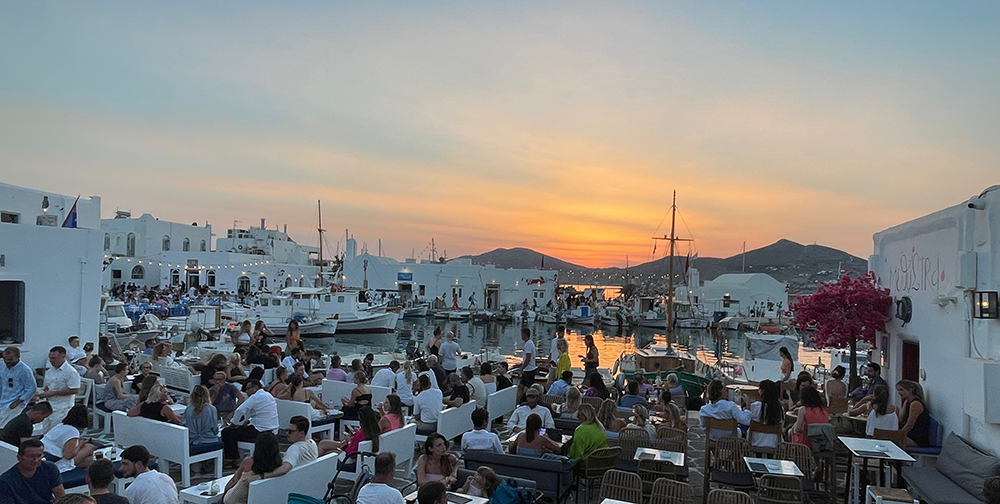 Paros Adası Naousa'daki Methystra barında, gün batımının sıcak turuncu tonlarına bürünmüş gökyüzü altında, limandaki beyaz binalar ve demirli tekneler eşliğinde, açık avludaki masalarda oturan kalabalıklar keyifli bir akşam geçiriyor, bu Paros seyahat rehberinin sunduğu huzurlu anlardan birini yansıtıyor.