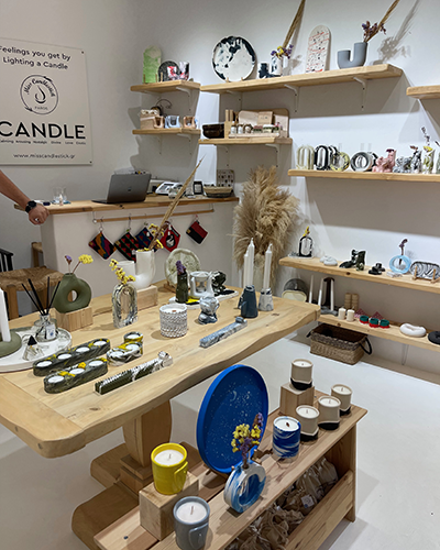 Paros seyahat rehberinin önerdiği Miss Candlestick mağazasında, geniş ahşap bir masa ve duvardaki raflar üzerinde sergilenen çeşitli el yapımı doğal mumlar, estetik oda kokuları ve sanatsal seramik objeler, Paros'un dinlendirici atmosferini yansıtan sıcak bir ortam sunuyor.