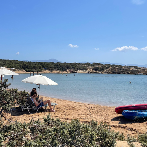 Paros seyahat rehberinin sakin plaj önerilerinden biri olan Paralia Santa Anna'nın huzurlu atmosferini yansıtan bu fotoğrafta, beyaz bir şemsiyenin altında dinlenen bir kişi ve berrak denizde serinleyen tatilcilerle çevrili, yemyeşil bitki örtüsüyle kaplı tepelerin ve uzak dağların eşlik ettiği doğal bir Paros plajı gözüküyor.