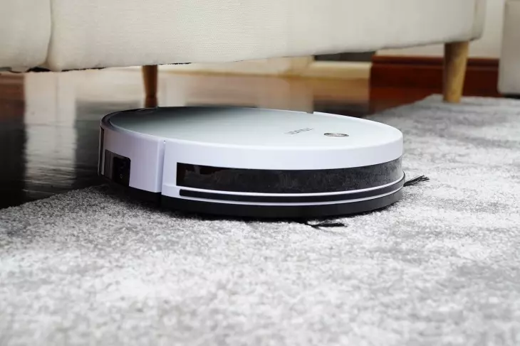 Beyaz robot süpürge, koltuk altına doğru ilerlerken gri halı üzerinde yakın açıdan görünüm.