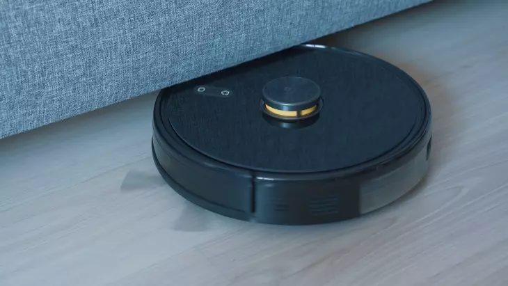 Siyah robot süpürge, koltuk altındaki dar alanda temizlik yaparken açık renk zemin üzerinde yakın çekim görünüm.