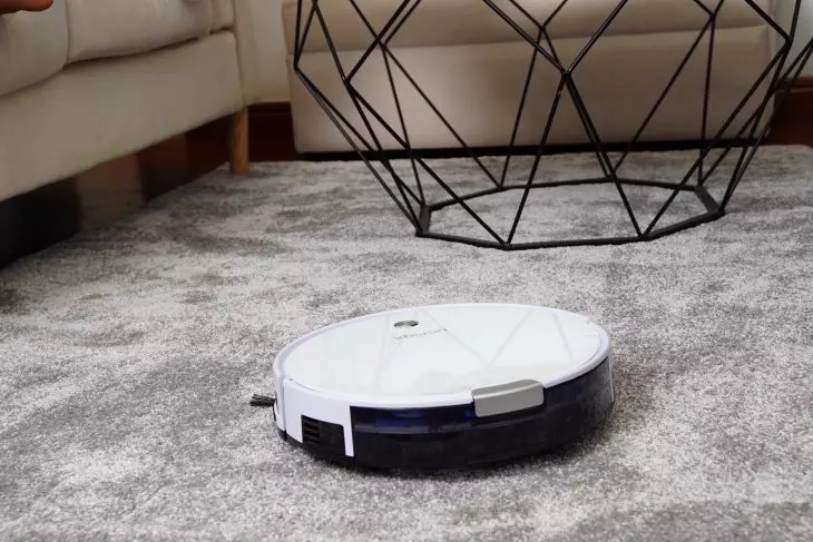 Beyaz robot süpürge, gri halı üzerinde temizlik yaparken arka planda modern metal ayaklı sehpa bulunan oturma odası görünümü.