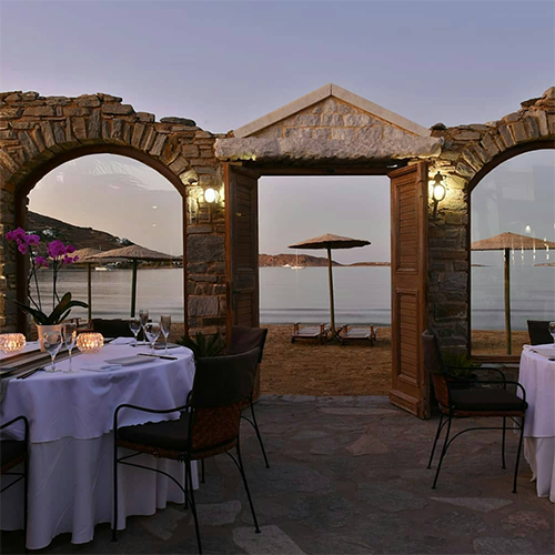 Paros Adası'nda, kumsala açılan taş kemerler ve kapı çerçevesiyle çevrili, masaları mor orkide ve mum ışıklarıyla süslenmiş şık bir açık hava restoranı, uzakta bir tekne, hasır şemsiyeler ve şezlongların seçildiği, gün batımının huzurlu atmosferini yansıtan bu kare, Astir of Paros gibi lüks bir tesiste sunulan dinlendirici bir tatil deneyimini vurguluyor.