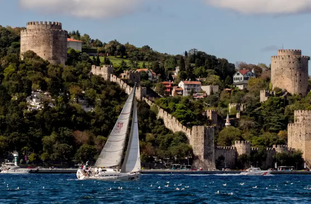 Beyaz yelkenli bir tekne, TUR 1937 numaralı yelkenleriyle Boğaz Kupası yarışında süzülürken, yemyeşil tepelerin üzerinde yükselen tarihi Rumeli Hisarı'nın taş duvarları ve kuleleri etkileyici bir fon oluşturuyor.