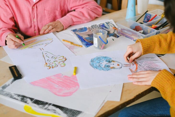 Ahşap bir masada oturan iki kişinin, biri pembe diğeri hardal sarısı kazaklı, farklı renklerde pastel boyalarla yüz çizimleri ve soyut şekillerin olduğu beyaz kağıtları boyarken, evden çalışırken yaratıcılığı ve motivasyonu artıracak mola ve ilham kaynakları bulmanın önemini vurgulayan samimi bir an.