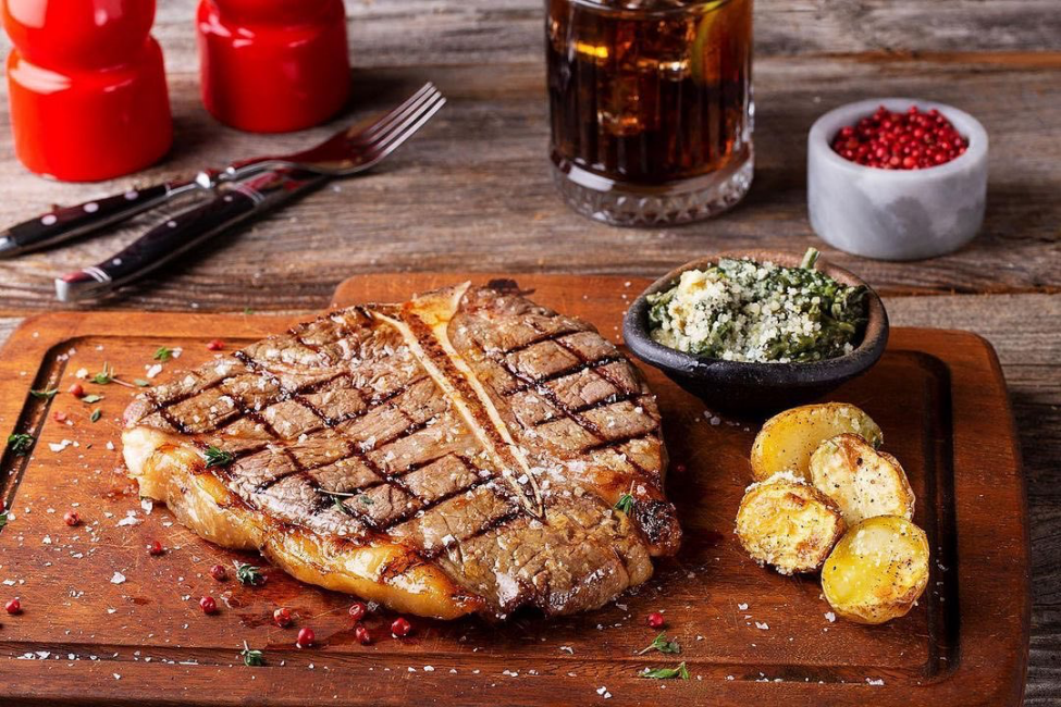 Tahta bir servis tahtası üzerinde ızgara izleriyle mükemmel pişmiş, üzerine deniz tuzu ve taze kekik serpilmiş T-bone steak, yanında parmesanlı ıspanak, kavrulmuş patatesler ve pembe karabiber taneleriyle servis edilerek İstanbul'un en iyi et restoranları arasında bir steakhouse deneyimini sunuyor.