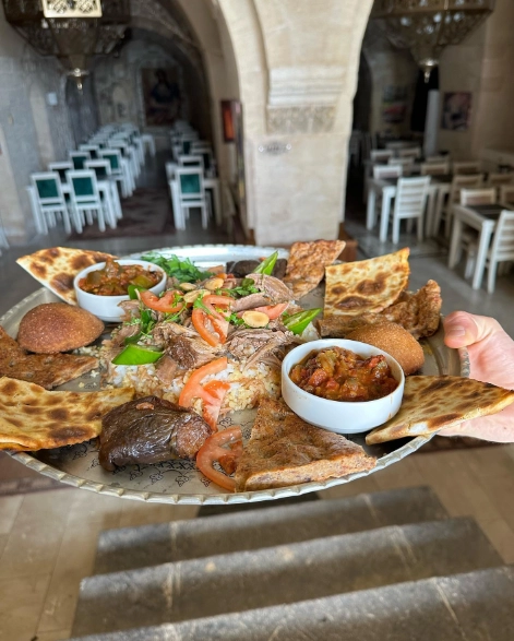 Büyük, gümüş bir tepside, ızgara karides, bol dilimlenmiş et, baharatlı pilav, pide ekmekleri ve iki kase meze ile donatılmış zengin bir sunum; arka planda yeşil ve beyaz sandalyelerin görüldüğü geleneksel bir restoran ambiyansında sunulan görkemli bir yemek ziyafeti.
