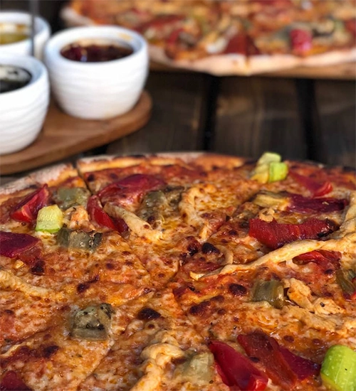 Yakın çekimde, sucuk, kırmızı ve yeşil biber dilimleri, bol erimiş peynir ile zengin bir şekilde süslenmiş, kenarları çıtırlaşmış bir bütün pizza ve arka planda ahşap bir zeminde duran, içinde soslar olan küçük kaseler ile flu başka bir pizza görülüyor.