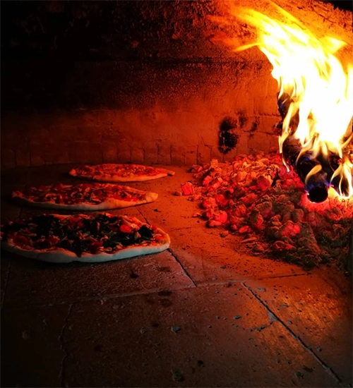 Geleneksel taş fırında, sağ tarafta yükselen parlak alevlerin yoğun ısısı altında, koyu renkli fırın zemininde üzeri bol malzemeli üç adet lezzetli pizza veya lahmacun pişerken, yiyeceklerin üzerindeki malzemeler ateşin kızıl ışığında parlıyor ve iştah açıcı bir atmosfer yaratıyor.