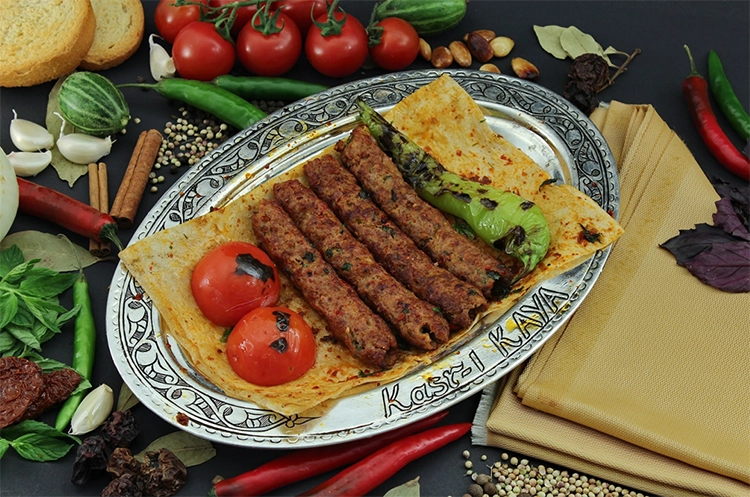 Şık gümüş bir tabağın merkezinde lavaş üzerinde beş adet iştah açıcı ızgara Adana kebap, iki közlenmiş domates ve bir közlenmiş yeşil biberle özenle sunulmuş; Kasr-ı Kaya ismini taşıyan bu tabak, arka planda taze domates, sarımsak, tarçın çubukları, karabiber taneleri, kuru domates, nane ve kırmızı biberler gibi çeşitli malzemelerle çevrili, zengin bir gastronomik şöleni gözler önüne seriyor.