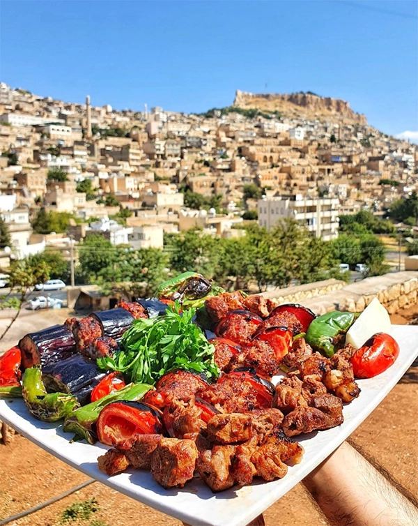 Mardin'in tarihi dokusunu yansıtan taş evleri ve zirvedeki kalesinin nefes kesen manzarası önünde, Adana kebap, şiş kebap, közlenmiş patlıcan, domates, biber ve taze yeşilliklerle bezenmiş bol çeşitli bir kebap tabağı sunuluyor.