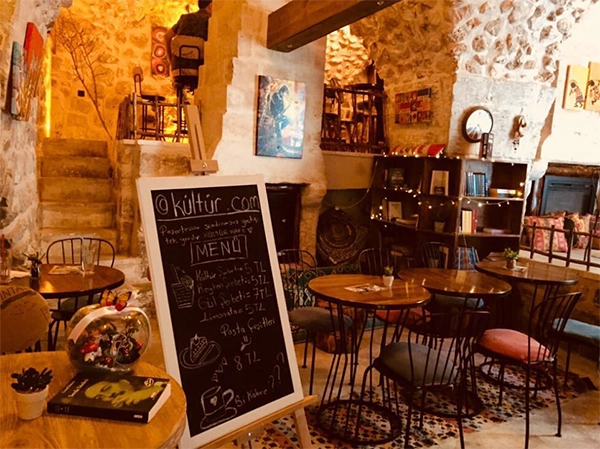 Taş duvarlara sahip, loş ışıklı, samimi bir kültür kafe iç mekanı; ön planda bir menü tahtası, arka planda yuvarlak masalar, sandalyeler, raflarda ve masalarda kitaplar ile duvarlarda sanat eserlerinin bulunduğu, huzurlu bir ortam sunuyor.