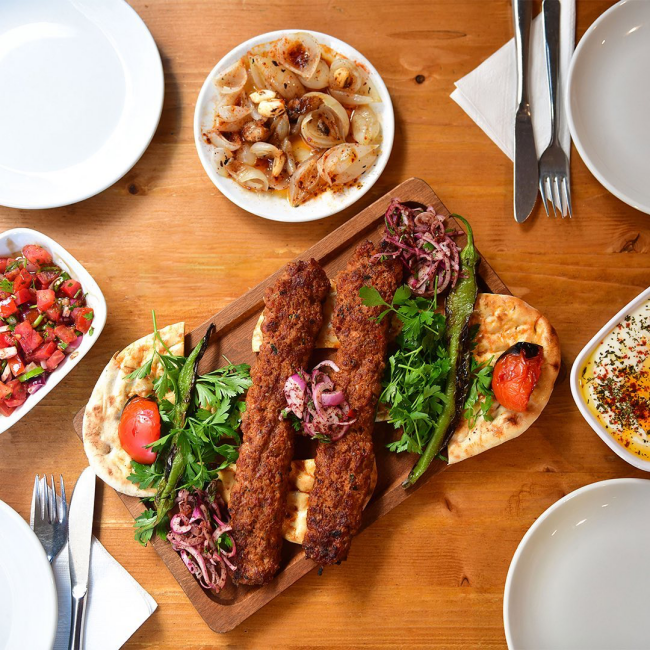 Bir tepsi üzerinde servis edilen, közlenmiş biber ve domates, sumaklı soğan ve taze maydanozla süslenmiş üç adet iştah açıcı Adana kebap, yanında gavurdağı salatası, tereyağlı soğan ve baharatlı yoğurtlu bir meze ile İstanbul'un en iyi et restoranları deneyimini yansıtan zengin bir kebap sofrası.