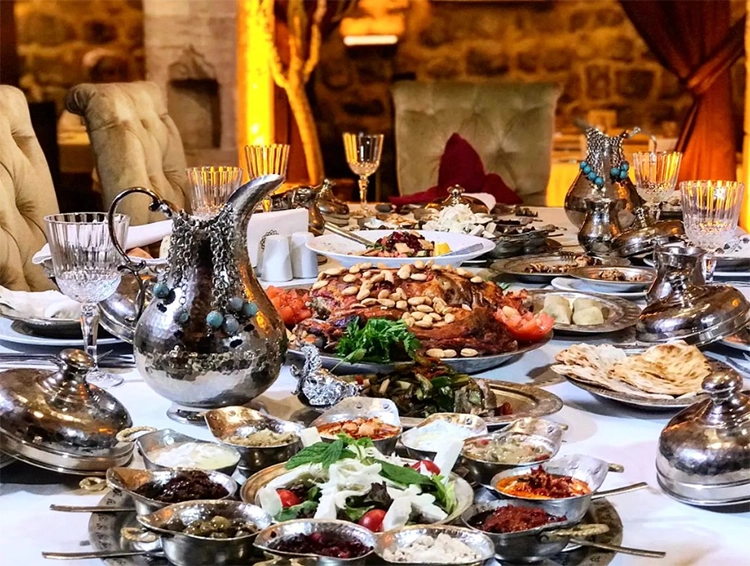 Arka planda taş duvarlar ve rahat sandalyelerin görüldüğü, zengin bir atmosferde, ön planda işlemeli gümüş bir sürahi, çok sayıda gümüş servis kasesi ve tabaklarının yanı sıra bademli ana yemek, bol çeşitli mezeler, taze yeşillik salatası ve lavaşların yer aldığı, geleneksel bir ziyafet sofrası.