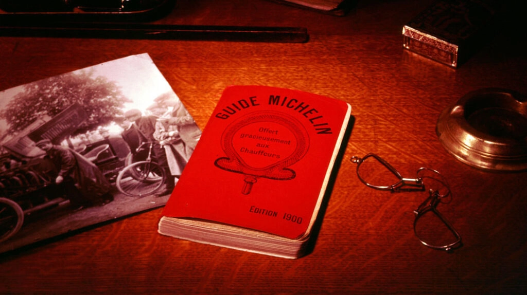 1900 yılına ait ilk Michelin Rehberi’nin kırmızı kapaklı baskısı, eski bir masa üzerinde gözlük ve siyah beyaz bir fotoğrafla birlikte sergileniyor.