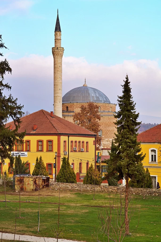 Üsküp'teki tarihi Mustafa Paşa Camisi'nin zarif minaresi ve gri kubbesi, önünde sarı renkli binalar, kozalaklı ağaçlar ve yeşil bir alanla çevrili olarak, Balkanlar'ın zengin kültürel mirasını gözler önüne seriyor.