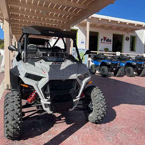 Beyaz renkli, geniş ve dişli lastikli bir ATV, kırmızı süspansiyon detaylarıyla dikkat çekerek, Paros adasını keşfetmek isteyenler için Ride Moto Rental dükkanının önünde, arkada sıralanmış mavi ATV'lerle birlikte, Paros Seyahat Rehberi'nde bahsedilen ada içi ulaşım seçeneklerini vurgulayarak park etmiş görünüyor.