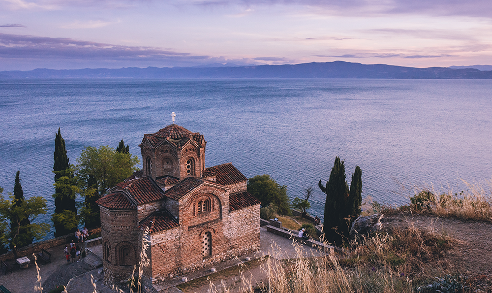 Ohrid Gölü kıyısında, gün batımının mor ve turuncu tonlarına bürünmüş gökyüzü altında, kendine özgü taş mimarisi ve kiremit çatısıyla öne çıkan, etrafı yeşil ağaçlarla çevrili tarihi Aziz Yuhanna Kilisesi, Üsküp seyahat rehberi kapsamında gezginleri tarihten kalan izleriyle buluşturan büyüleyici bir durak sunuyor.