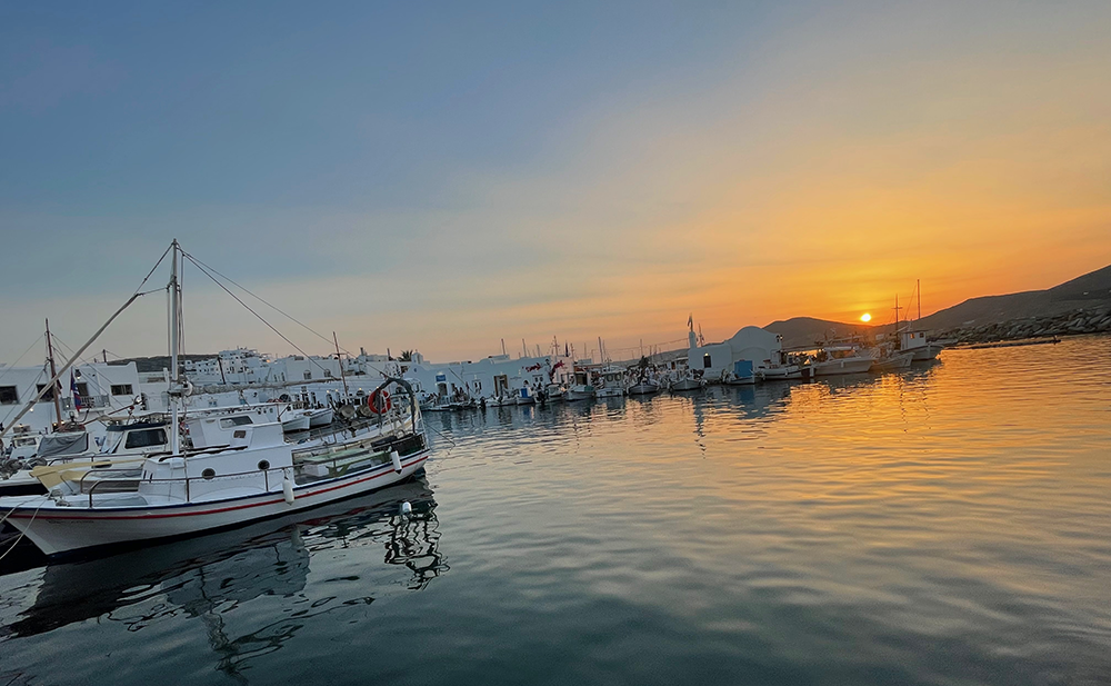 Paros Adası'nda, gün batımının turuncu tonlarıyla aydınlanan gökyüzünün sakin deniz suyunda yansıdığı, geleneksel beyaz binaların kuşattığı bir limanda demirli çok sayıda küçük balıkçı teknesi, adanın huzurlu ambiyansını gözler önüne seriyor.