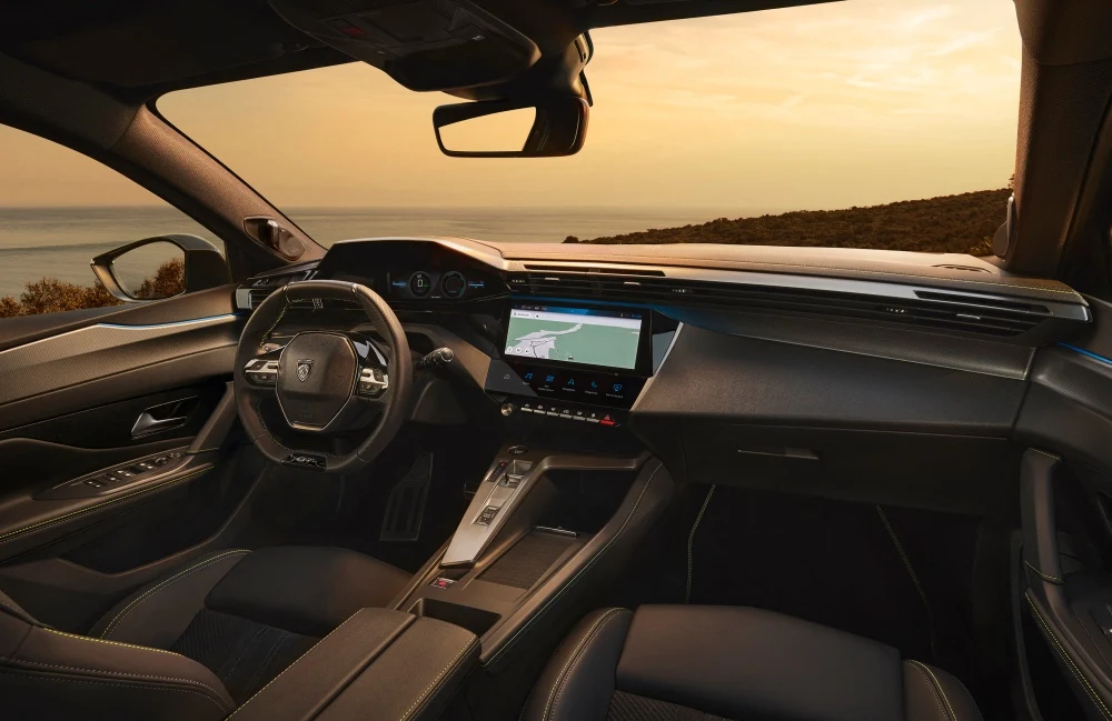 Peugeot 408 facelift modelinin iç mekanı, dijital gösterge paneli ve navigasyon haritası açık merkezi bilgi-eğlence ekranı ile modern i-Cockpit tasarımını gösterirken, ön camdan gün batımında engin bir deniz manzarası görünüyor.
