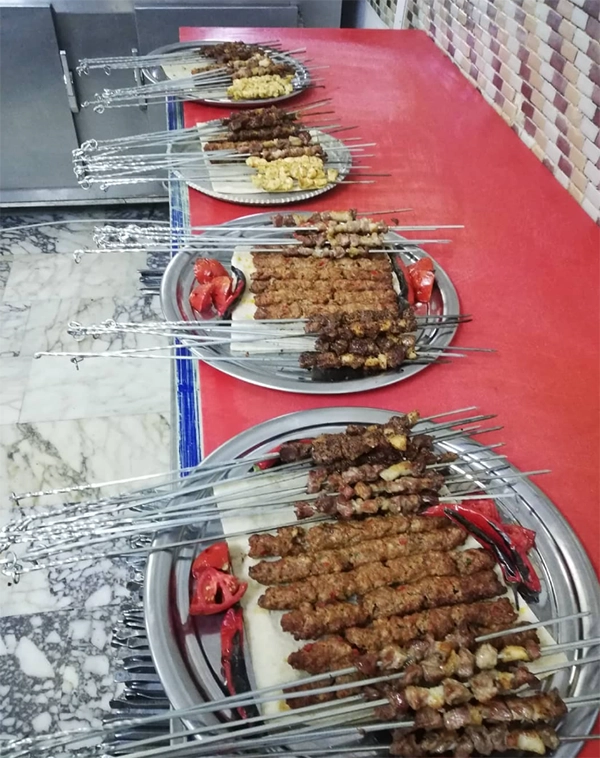 Kırmızı bir masa üzerinde yan yana dizilmiş metal tepsilerde, lavaş ekmeği üzerinde sunulan bolca Adana kebap, kuşbaşı et ve tavuk şiş kebap çeşitleri ile ızgara domates ve biberler, servise hazır bir şekilde bekliyor.